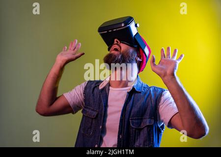 Beeindruckender Hipster-Typ mit Virtual-Reality-Brille hob seine Hände hoch und bekommt ein neues digitales Erlebnis, der aufschaut und Jeans-Weste trägt. Innenaufnahmen im Studio, isoliert auf farbigem Neonlicht-Hintergrund. Stockfoto