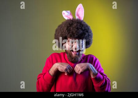 Staunend aufgeregt Hipster Mann mit Afro-Frisur und Hasenohren, Pfoten mit seinen Händen, Blick auf die Kamera, trägt rote Sweatshirt. Innenaufnahmen im Studio, isoliert auf farbigem Neonlicht-Hintergrund. Stockfoto