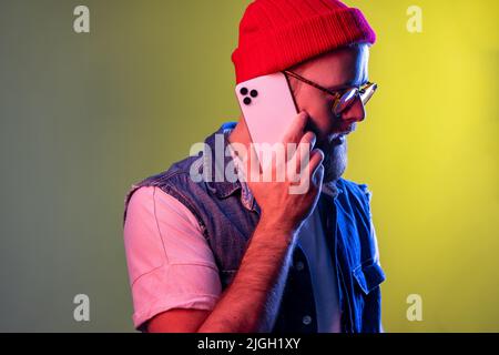 Seitenansicht des jungen erwachsenen Hipster-Mannes, der telefonieren konnte, mit ernstem konzentriertem Gesichtsausdruck, mit Beanie-Hut und Denim-Weste. Innenaufnahmen im Studio, isoliert auf farbigem Neonlicht-Hintergrund. Stockfoto