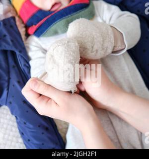 Eine Mutter legt warme Fäustlinge für einen kleinen Jungen an. Mama kleidet ein glückliches Kind in Winterkleidung auf dem Bett. Stockfoto
