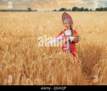 Kleines Mädchen im russischen nationalen Sarafan und ein Kokoschnik, der am Sommertag in einem goldenen Weizenfeld steht Stockfoto