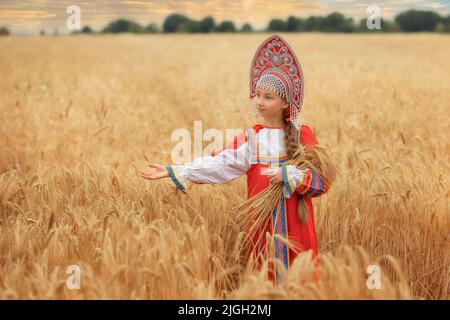 Kleines Mädchen im russischen nationalen Sarafan und ein Kokoschnik, der am Sommertag in einem goldenen Weizenfeld steht Stockfoto