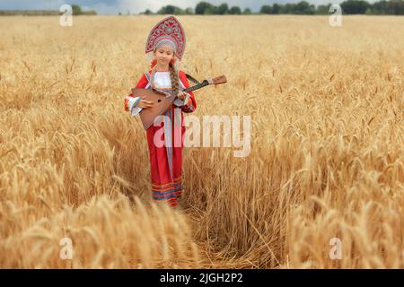 Kleines Mädchen im russischen nationalen Sarafan und ein Kokoschnik, der am Sommertag in einem goldenen Weizenfeld steht Stockfoto