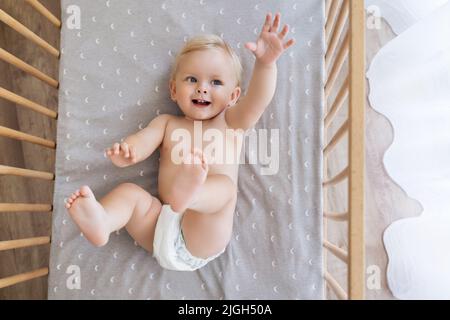 Blick von oben auf den kleinen verspielten Jungen in der Windel, der auf dem Rücken liegt und die Hand ausstreckt, um Spielzeug oder Werbetexte zu berühren und in seinem Bett zu spielen. Konzept für die Kinderbetreuung. Frohe Kindheit Stockfoto