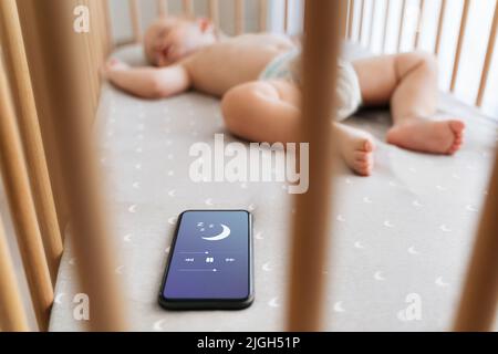 Selektiver Fokus des kleinen Jungen in der Krippe Einschlafen mit Hilfe der Anwendung für Babyschlaf am Telefon neben ihm liegen, während Mutter Abendessen in der Küche kochen. Kinderbetreuung und Technologie Stockfoto