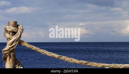 Altes Seil an Holzpfosten am Meer geknüpft, blaues Meer und wolkiger Himmel an sonnigen Sommertagen. Panoramablick. Stockfoto