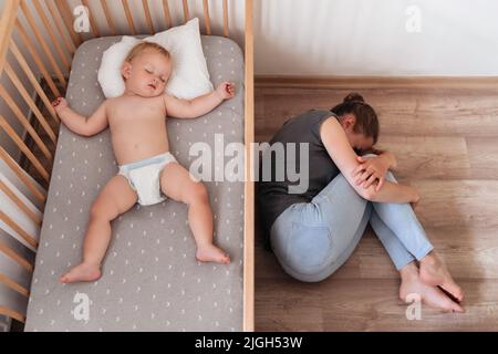 Obere Ansicht eines niedlichen Babys, das in seinem Bett neben seiner Mutter, die weinend auf dem Boden liegt, verzweifelt und einsam ist und an postnataler Depression leidet. Schwierigkeiten der Mutterschaft Stockfoto