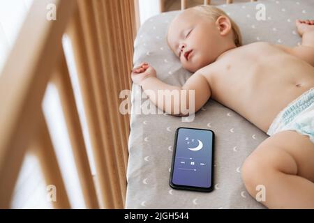 Kleines Kind in Windelnaben im Bett mit Telefon spielen Schlaflied in Anwendung für Baby schlafen im Schlafzimmer, während die Eltern ruhen im Wohnzimmer. Glückliche Mutterschaft und Technologie Stockfoto