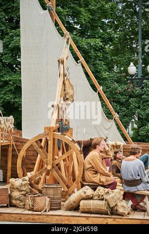 Menschen in den Kleidern des Mittelalters sitzen auf einem alten Pier in der Nähe eines alten Segelbootes. Times and Epochs Festival auf den Straßen von Moskau, Russi Stockfoto