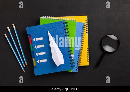 Das Konzept von Bildung und Zukunftsplanung. Nahaufnahme eines Stapels von farbigen Heften, einer Lupe, Bleistiften und einem Papierflugzeug - ein Symbol für Stockfoto