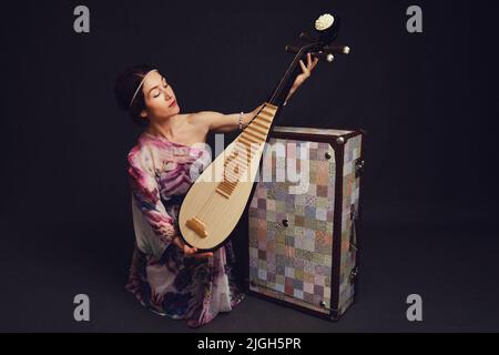 Eine glückliche Musikerin in asiatischen Kleidern auf einem schwarzen Hintergrund im Studio. Eine lächelnde Frau mit einem Saiteninstrument aus Asien auf einem dunklen Backgrou Stockfoto