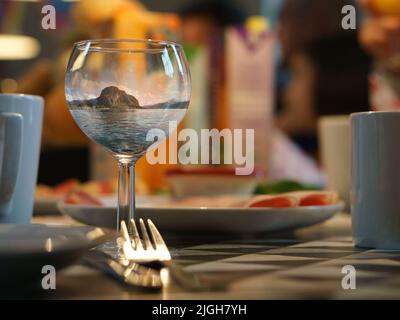 Fantasy-Bild, das aus einem Weinglas mit der Landschaft aus Norwegen am Meer im Glas komponiert. Auf einem Esstisch mit Besteck, Tellern und Tassen. Creat Stockfoto