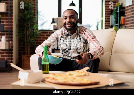 Junge Erwachsene wechseln den fernsehkanal mit Fernbedienung und essen Chips, finden Film auf dem Fernseher zu Hause zu sehen. Genießen Sie Snacks und Bierflaschen mit Lieferservice zum Mitnehmen auf dem Sofa. Stockfoto