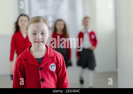 Fröhliche Schulkameradinnen mit Down-Syndrom in Uniform, die im Schulkorridor mit Klassenkameraden hinter ihr spazieren gehen. Stockfoto