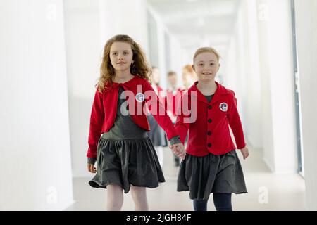 Fröhliche Schulkameradin mit Down-Syndrom in Uniform, die ihre Hände hält und ihre Klassenkameradin im Schulkorridor mit Klassenkameraden hinter sich herumläuft. Stockfoto