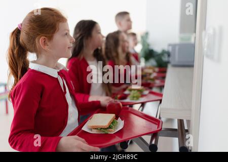 Fröhliche Schulkinder, die mit Tabletts in der Schlange stehen und in der Schulkantine zu Mittag essen. Stockfoto