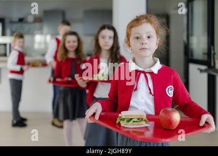 Glückliche Schüler in Uniformen, die Tabletts mit Mittagessen in der Warteschlange in der Schulkantine halten. Stockfoto