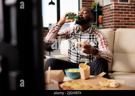 Junger Erwachsener trinkt alkoholisches Bier aus der Flasche und isst Essen zum Mitnehmen aus dem Fast-Food-Lieferpaket, schaut sich einen Film im fernsehen an. Genießen Sie einen Drink und eine Mahlzeit zum Mitnehmen vor dem Fernseher. Stockfoto