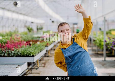 Junge Angestellte mit Down-Syndrom, die im Gartencenter arbeitet, die Kamera anschaut und winkt. Stockfoto