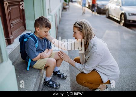Mutter tröstete ihren kleinen Sohn an seinem ersten Schultag, saß auf einer Treppe und sagte vor der Schule Auf Wiedersehen. Stockfoto