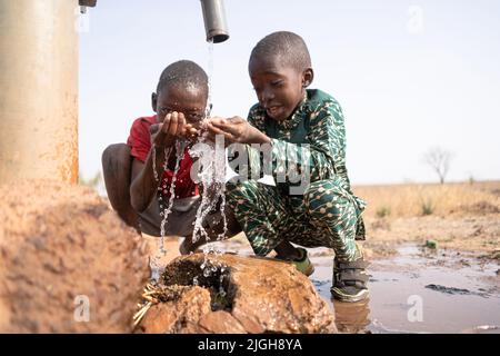 Zwei glückliche Jungen, die mit frischem Wasser aus einem Dorfhahn in einer afrikanischen Steppenregion spielen; Desertifikationsfortschritt und Wasserknappheitskonzept Stockfoto