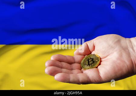 Goldene Bitcoin-Münze in der Hand des Mannes, ukrainische Flagge im Hintergrund. Stockfoto