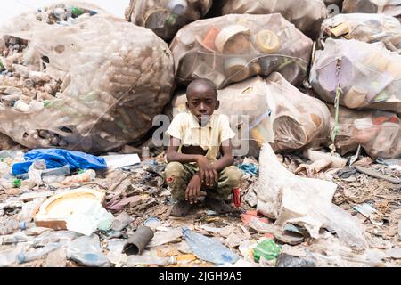 Junger Africanboy sitzt vor riesigen Tüten voller sortierter recycelbarer Plastikflaschen auf einer Mülldeponie; rudimentäre Abfallwirtschaft in der Entwicklung Stockfoto