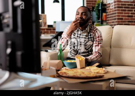 afroamerikanischer Mann, der Chips isst, während er sich einen Film im fernsehen ansieht und Spaß mit der Lieferung zum Mitnehmen und einer Bierflasche hat. Sehen Sie sich einen Film im Fernsehen an und genießen Sie zu Hause ein Fastfood-Essen zum Mitnehmen. Stockfoto