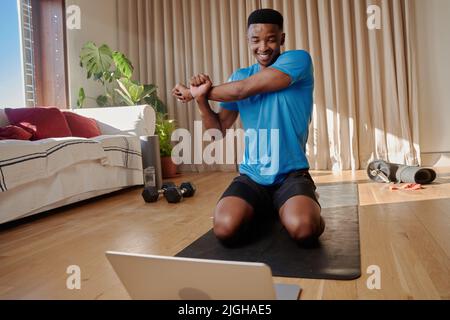 Der junge schwarze Afroamerikaner arbeitet glücklich zu Hause und streckt sich die Arme, bevor er auf seiner Yogamatte in seinem Modern einen Online-Trainingskurs macht Stockfoto
