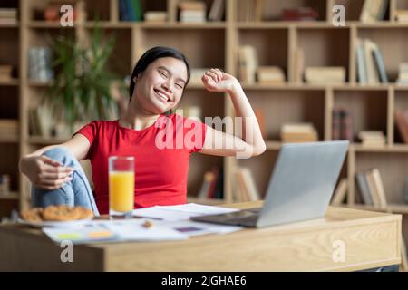 Zufriedene glückliche junge chinesische Frau mit Down-Syndrom streckt den Körper und ruht sich mit Laptop am Tisch aus Stockfoto