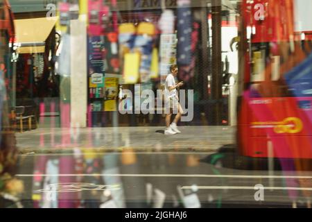 Ein Mann geht die neue Oxford Street entlang und schaut auf sein Telefon. Durch das Schaufenster des Regenschirms geführt. Reflexionen.künstlerisch. London Bus. Schwenken. Kreativ. Stockfoto