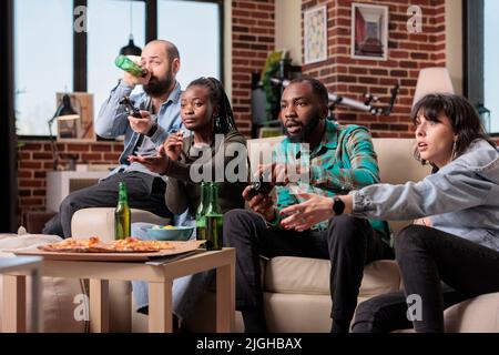Eine Gruppe verschiedener Freunde, die zusammen Videospiele spielen und sich zu Hause mit Bier und Essen treffen. Genießen Sie Spielwettbewerbe auf der Fernsehkonsole, jubelnde und trinkende Menschen. Stockfoto