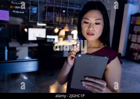 Serious asiatische Geschäftsfrau mit digitalen Tablet beim Schreiben auf Glaswand im Büro in der Nacht Stockfoto