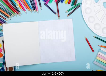 Leeres Blatt Papier eines offenen mathematischen Notizbuchs, Kunstschulmaterialien für Malerei, auf blauem Hintergrund mit Platz für Text. Bunte Bleistifte, markieren Stockfoto