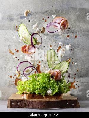 Sandwich mit fliegenden Zutaten auf Holzbrett auf texturiertem Hintergrund. Levitation gesunde Ernährung kreatives Konzept. Vertikale Ausrichtung Stockfoto
