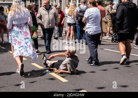 Ein Mann, der auf der Straße liegt, während Menschen in Penzance in Cornwall, England, an ihm vorbeigehen. Stockfoto