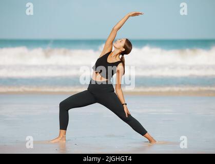 Streckt und stärkt euren Weg zu einem besseren Körper. Eine junge Frau macht einen Halbmondschritt, während sie am Strand Yoga praktiziert. Stockfoto
