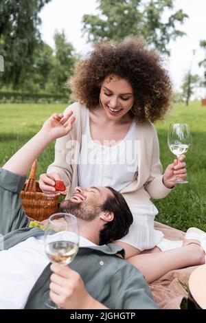 Fröhliche lockige Frau mit einem Glas Wein und Fütterung Freund mit frischen Erdbeeren während des Picknicks Stockfoto