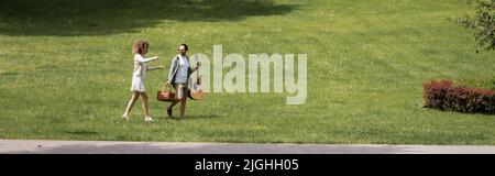 In voller Länge Mann mit Gitarre und Weidenkorb in der Nähe glückliche Freundin gestikulieren in grünen Park, Banner Stockfoto