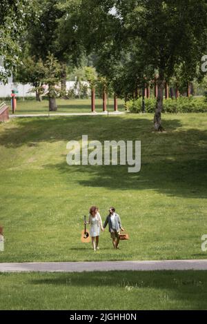 Die ganze Länge des Paares in Sommerkleidung trägt Gitarre und Picknick Korbkorb, während Sie im grünen Park spazieren Stockfoto