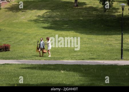 Die ganze Länge des Paares in Sommerkleidung trägt Gitarre und Picknick Weidenkorb im grünen Park Stockfoto