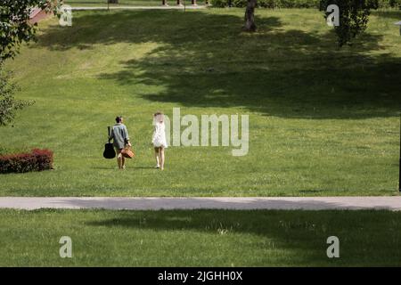 Volle Länge der Mann in Sommerkleidung tragen Gitarre und Picknick Weidenkorb in der Nähe Freundin im grünen Park Stockfoto