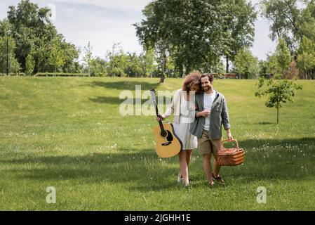 Die ganze Länge der glücklichen lockigen Frau hält akustische Gitarre in der Nähe des Mannes, der Korbkorb trägt, während er im grünen Park geht Stockfoto