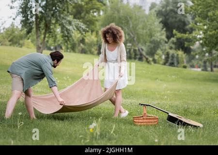 Fröhliches junges Paar in Sommerkleidung Putting Picknick Decke auf grünem Gras im Park Stockfoto