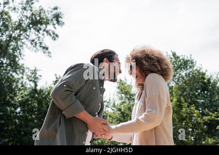 Low-Angle-Ansicht des fröhlichen Mannes und der glücklichen lockigen Frau in einer trendigen Sonnenbrille, die die Hände im Park hält Stockfoto
