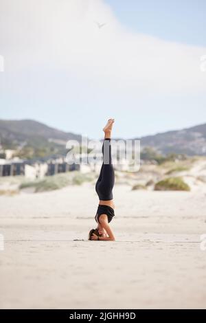 Es braucht Geduld, um diese Pose zu meistern. Eine junge Frau, die einen Kopfstand macht, während sie am Strand Yoga praktiziert. Stockfoto