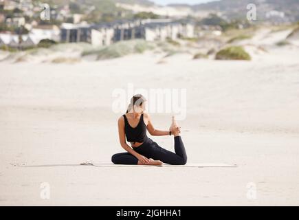 Das Meer bildet die perfekte Landschaft, um an sich selbst zu arbeiten. Eine junge Frau, die eine Taubenpose macht, während sie am Strand Yoga praktiziert. Stockfoto