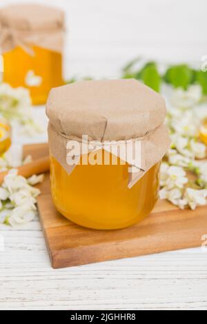 Süßes Honigglas umgab die Akazienblüten des Frühlings. Honig fließt aus einem Löffel in ein Glas. Gläser von klarem frischen Akazienhonig auf Holzgrund. Stockfoto