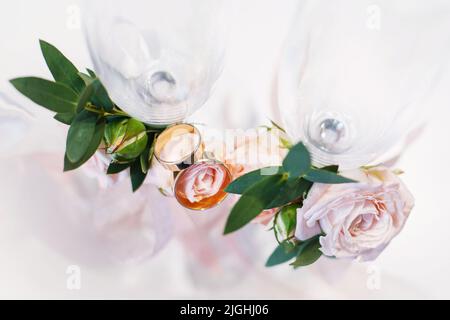 Hochzeitsdetails. Zwei goldene Eheringe aus nächster Nähe liegen auf Blumen, die Champagnergläser schmücken. Hochzeitsdetails, Symbol für Liebe und Ehe. Konzept von Stockfoto
