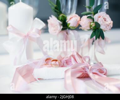 Zwei goldene Eheringe liegen auf dem Tisch neben den Hochzeitszubehör. Die Eheringe liegen auf dem Tisch neben den Kerzen mit rosa Bändern und Gläsern Stockfoto